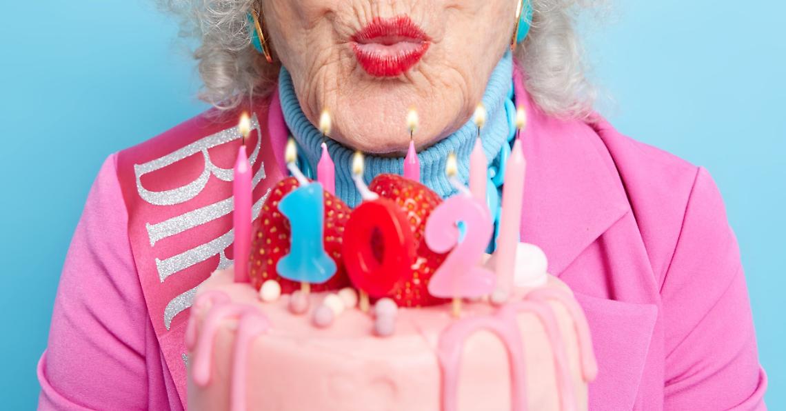 Close up shot of senior woman with grey culry hair keeps lips folded going to blow candles on cake celebrates birthday being well dressed has bright makeup enjoys special occasion gets congratulations