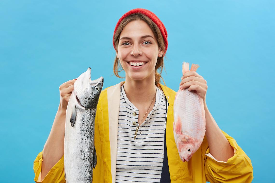 Successful female angler holding two big fish in hands, smiling at camera while having good mood after huge catch at river, posing against blue blank wall. Pretty fisherwoman with cheerful expression